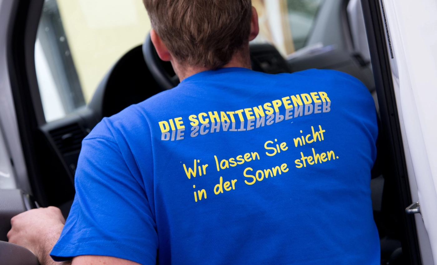 Mann, der ein T-Shirt mit der Aufschrift "Die Schattenspender - Wir lassen Sie nicht in der Sonne stehen." steht